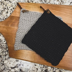 Op de afbeelding: Twee gebreide ovenwanten, een grijs en een zwart, met leren lussen en messing accenten. De ovenwanten liggen op een houten snijplank.