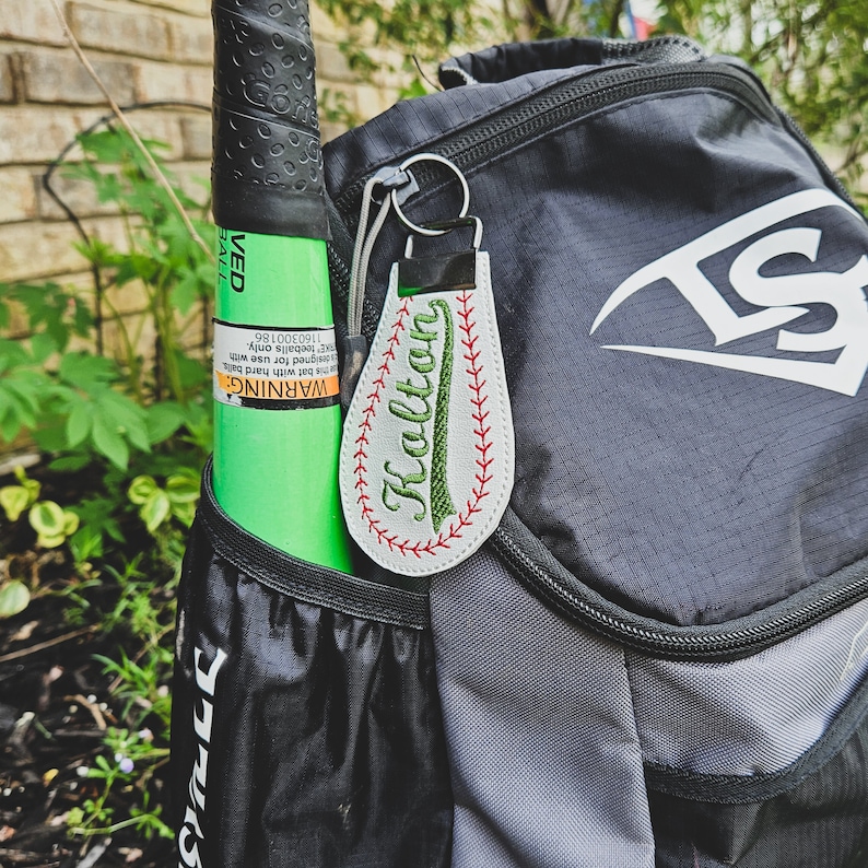 May include: A white baseball-shaped keyring with red stitching and a green embroidered name "Kalton" on a grey and black rucksack.