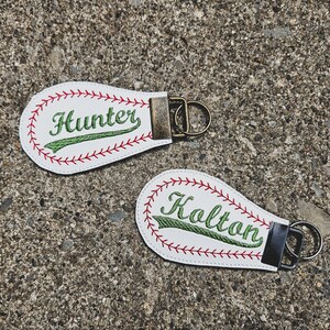 May include: Two white leather baseball-shaped keychains with red stitching. The keychains have green embroidered text, one says "Hunter" and the other says "Kalton".