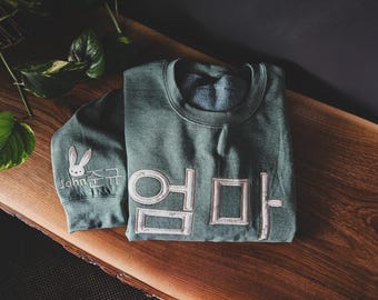 Puede incluir: Una sudadera doblada, de color verde azulado, con la palabra coreana "엄마" bordada en blanco. La manga presenta un conejito blanco y el nombre "John" en escritura cursiva. La sudadera está sobre una superficie de madera.