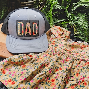 Puede incluir: Un gorro de camionero gris y negro con un parche floral que dice "DAD". El gorro está encima de un vestido floral amarillo.