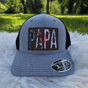 May include: Gray and black trucker hat with a brown patch that says "PAPA" in colorful patterned letters.