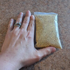 May include: A small, rectangular, clear plastic resealable bag filled with a coarse, golden-brown granular substance. The contents appear to be a dry spice blend or seasoning mix with visible flecks of varying textures and shades of tan and brown.