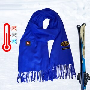 May include: A royal blue cashmere scarf with fringe, featuring a B-Vito logo and a heating element with a control panel. The scarf is displayed against a snowy background with a pair of skis.