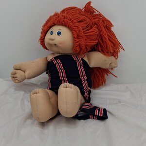 May include: A vintage doll with a soft, fabric body and a vinyl head. The doll has bright blue eyes, a rosy complexion, and a mass of orange yarn hair. It wears a navy blue and red striped outfit.