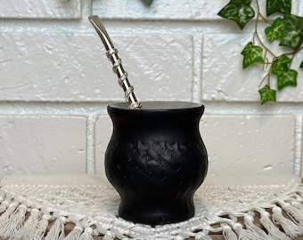 May include: Black ceramic mate gourd with a silver bombilla straw, sitting on a white crocheted doily.