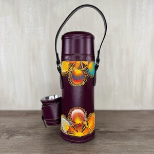 May include: A plum-coloured leather wine bottle carrier with a black handle and a matching cup holder. The carrier is adorned with colourful embroidered floral designs in yellow, orange, and blue. The carrier is designed to hold a standard-sized wine bottle.