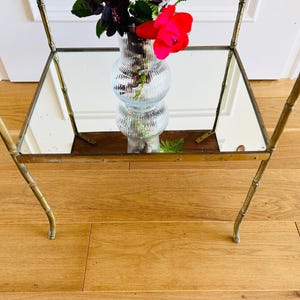 May include: A gold metal side table with a mirrored top and bamboo-style legs. A clear glass vase with pink flowers sits on the table.
