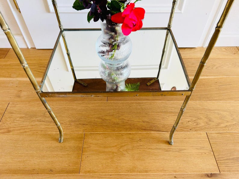 May include: A gold metal side table with a mirrored top and bamboo-style legs. A clear glass vase with pink flowers sits on the table.