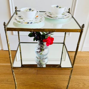 May include: A brass and mirrored two-tiered serving cart with a floral design on the top tier. Two teacups and saucers are on the top tier, and a vase with a single red rose is on the lower tier.