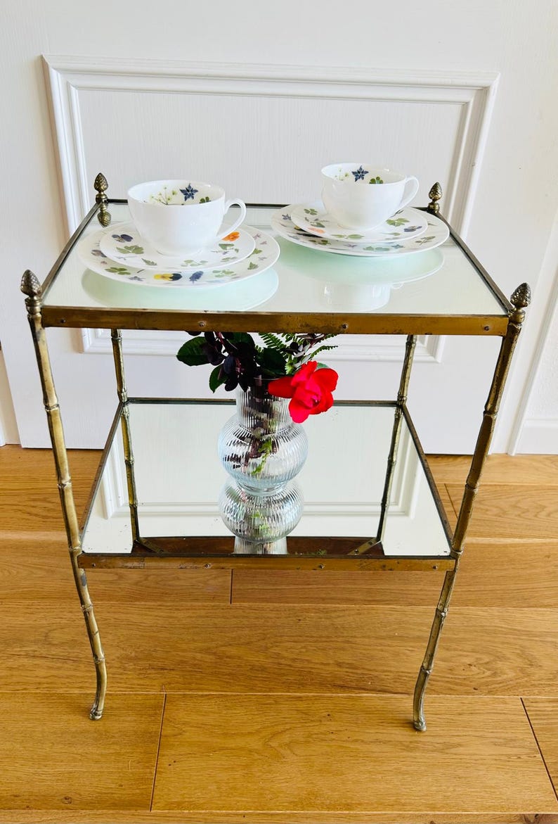 May include: A brass and mirrored two-tiered serving cart with a floral design on the top tier. Two teacups and saucers are on the top tier, and a vase with a single red rose is on the lower tier.
