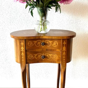 Può includere: Un tavolino da salotto in legno con due cassetti e dettagli intricati di intarsio. Il tavolo ha un piano curvo e tre gambe. Un vaso di peonie rosa è posto sopra il tavolo.