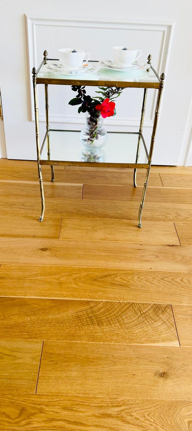 May include: A gold metal two-tiered serving cart with mirrored shelves. The cart has a floral arrangement with red flowers in a clear glass vase on the lower shelf. Two white teacups with floral patterns are on the top shelf.