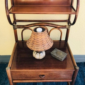 Puede incluir: Una mesita de noche de madera con un cajón y un estante. La mesita de noche tiene una lámpara de mimbre con una pantalla blanca en la parte superior. Una pequeña caja de plata está en el estante superior.
