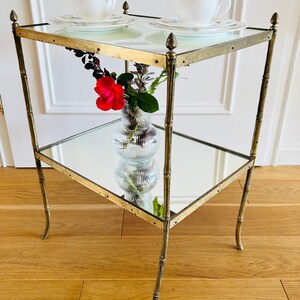 May include: A two-tiered brass side table with mirrored shelves. The table has a gold finish and features decorative finials on the top corners. A clear glass vase with a red rose and greenery sits on the lower shelf.