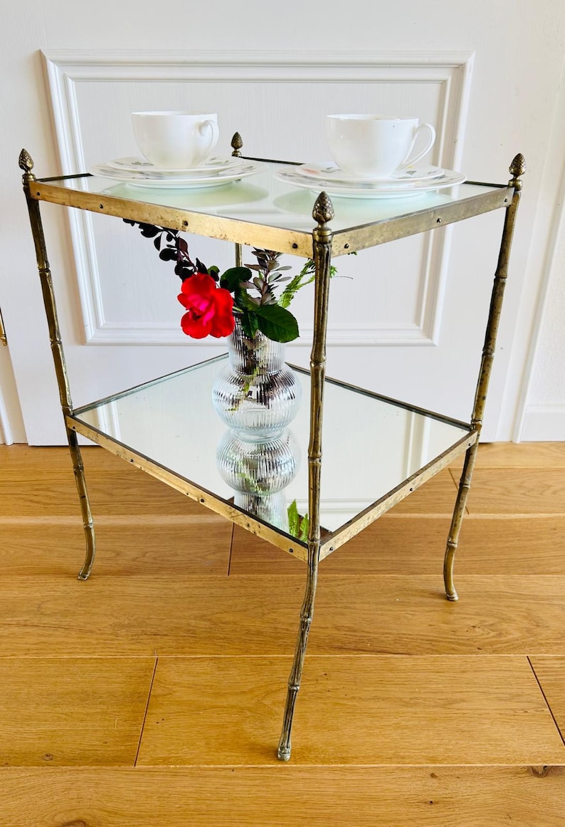 May include: A two-tiered brass side table with mirrored shelves. The table has a gold finish and features decorative finials on the top corners. A clear glass vase with a red rose and greenery sits on the lower shelf.