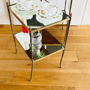 May include: Two mirrored glass-top tables with gold metal frames and legs. The tables are stacked on top of each other and have a floral patterned teacup and saucer on the top table.