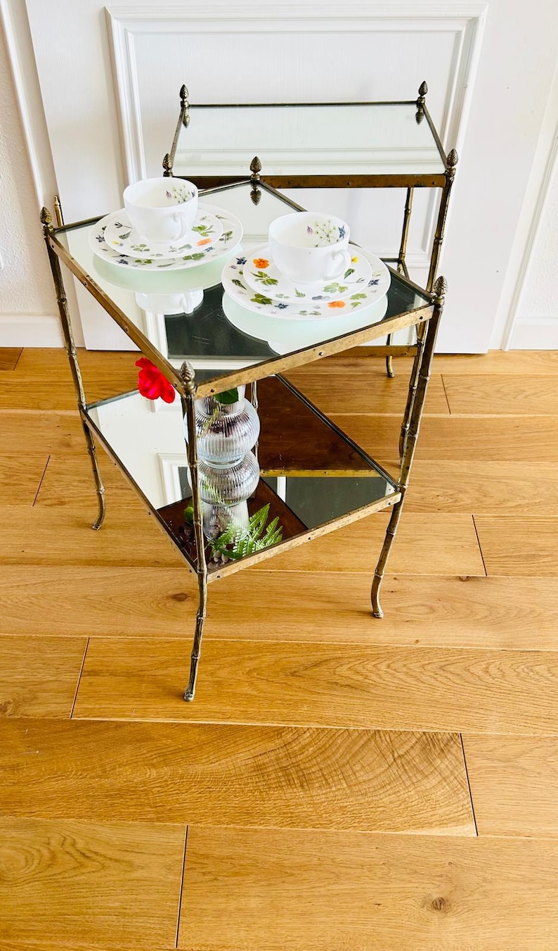 May include: Two mirrored glass-top tables with gold metal frames and legs. The tables are stacked on top of each other and have a floral patterned teacup and saucer on the top table.