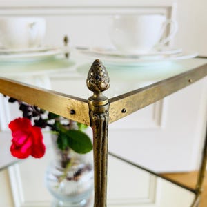 May include: A gold metal serving cart with glass shelves and a pine cone finial on each corner. The cart has a mirrored surface and is perfect for displaying decorative items or serving drinks.