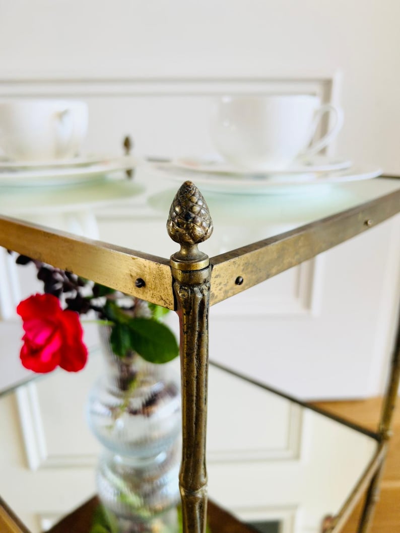 May include: A gold metal serving cart with glass shelves and a pine cone finial on each corner. The cart has a mirrored surface and is perfect for displaying decorative items or serving drinks.