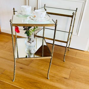 May include: Two mirrored side tables with gold metal frames and glass shelves. The tables are on a wooden floor.