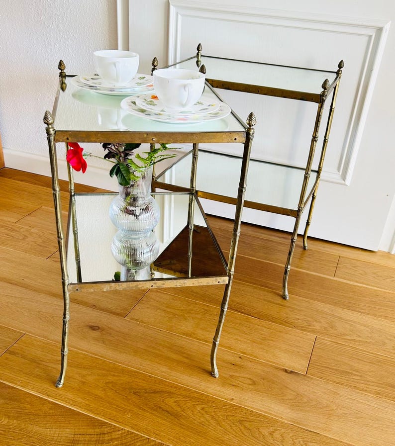 May include: Two mirrored side tables with gold metal frames and glass shelves. The tables are on a wooden floor.
