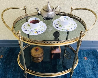 Puede incluir: Un carrito de té vintage de latón con dos estantes de vidrio. El estante superior contiene una tetera plateada, dos tazas de té con platillos y una taza de té. Los platillos y las tazas tienen un diseño floral. El estante inferior tiene una pequeña caja de madera y una botella decorativa.