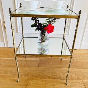 May include: A gold metal two-tiered table with mirrored shelves. The table has a bamboo-style design and features decorative finials on the top and bottom. A clear glass vase with a red rose and green foliage sits on the lower shelf.