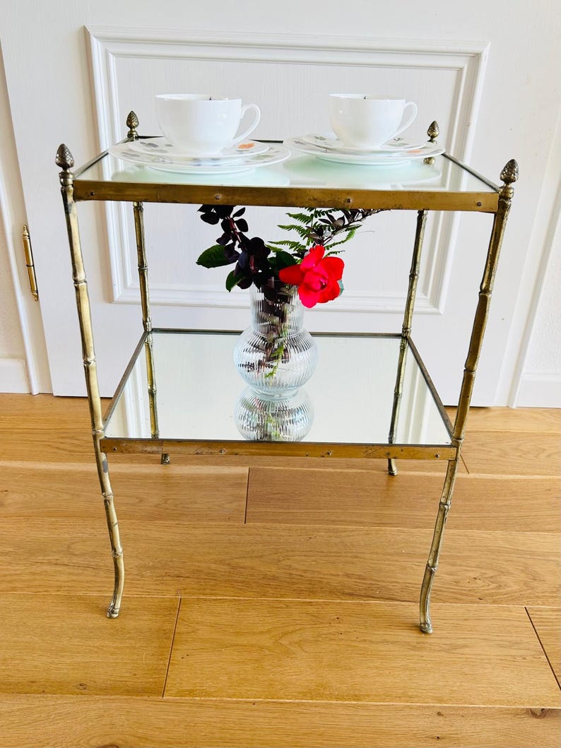 May include: A gold metal two-tiered table with mirrored shelves. The table has a bamboo-style design and features decorative finials on the top and bottom. A clear glass vase with a red rose and green foliage sits on the lower shelf.