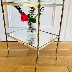May include: A gold metal side table with two glass shelves. The table has a bamboo-style design and features decorative accents. A vase with a red rose and greenery sits on the lower shelf.
