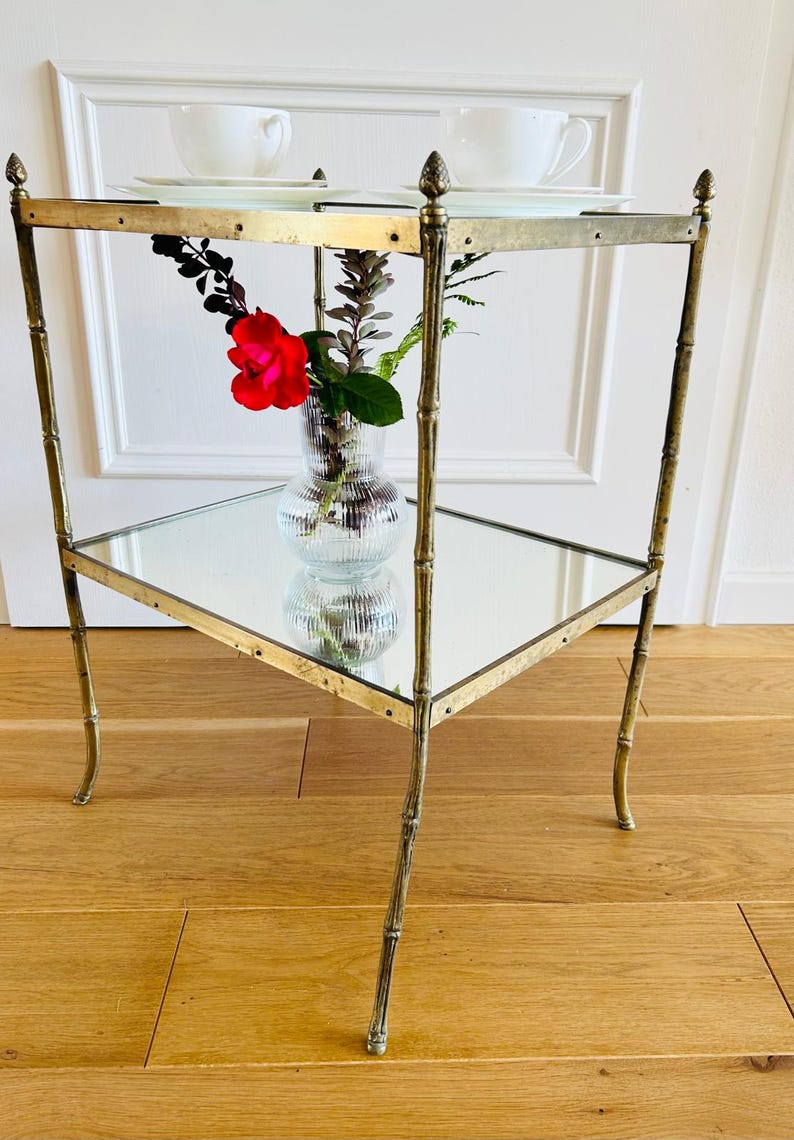 May include: A gold metal side table with two glass shelves. The table has a bamboo-style design and features decorative accents. A vase with a red rose and greenery sits on the lower shelf.