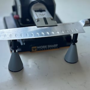 Może przedstawiać: Zbliżenie na nóż ostrzony na ostrzałce Work Sharp. Nóż spoczywa na dwóch szarych stożkach przymocowanych do ostrzałki. Ostrzałka jest czarna z żółtymi akcentami, a widoczny jest napis "Work Sharp".