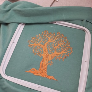 Könnte beinhalten: Ein orangefarbenes Baumdesign auf türkisfarbenem Stoff, gehalten in einem weißen Stickrahmen. Eine Spule beigefarbenes Garn und ein kleines Werkzeug sind im Hintergrund zu sehen, was auf ein Bastelprojekt hindeutet.