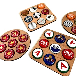 May include: Three wooden coasters with different designs. Each coaster has a circular or square shape and features a variety of colorful bottle caps arranged in a grid pattern. The bottle caps have different logos and designs, including beer brands, flags, and other symbols.