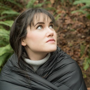May include: A person wearing a black puffer jacket, looking up, with a gray turtleneck underneath. The person is sitting in a forest setting with green ferns and brown leaves on the ground.
