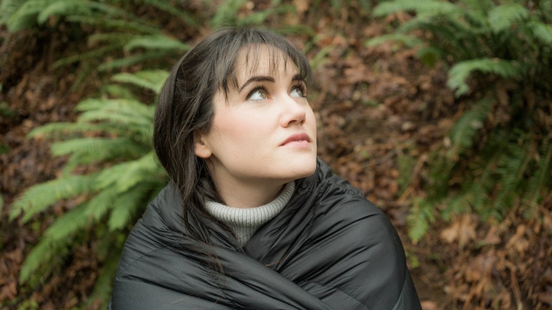 May include: A person wearing a black puffer jacket, looking up, with a gray turtleneck underneath. The person is sitting in a forest setting with green ferns and brown leaves on the ground.