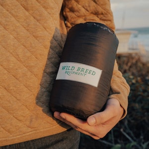 May include: A person holding a black sleeping bag with the text "WILD BREED EQUIPMENT" on a white label.