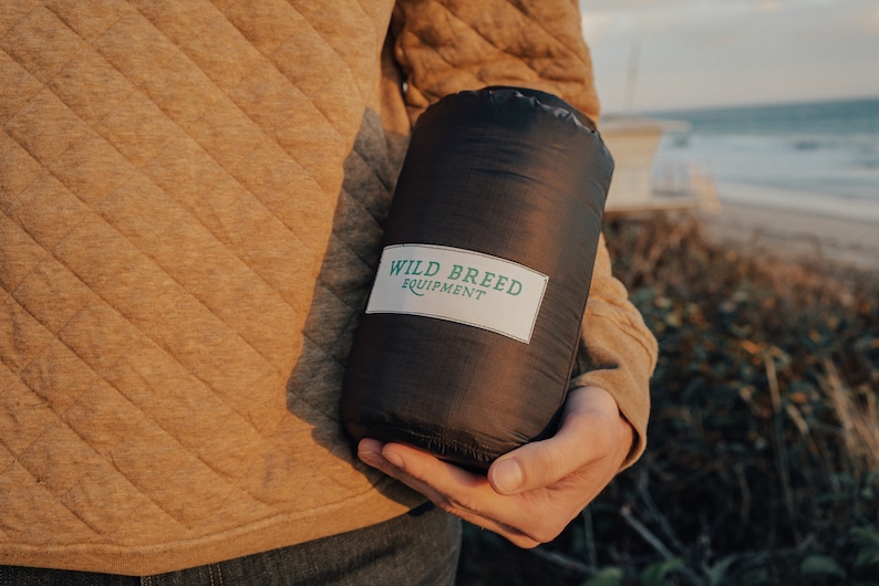 May include: A person holding a black sleeping bag with the text "WILD BREED EQUIPMENT" on a white label.