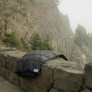 May include: A black down comforter is draped over a stone wall. The wall is on the edge of a road with a steep cliff in the background. The scene is foggy and misty.