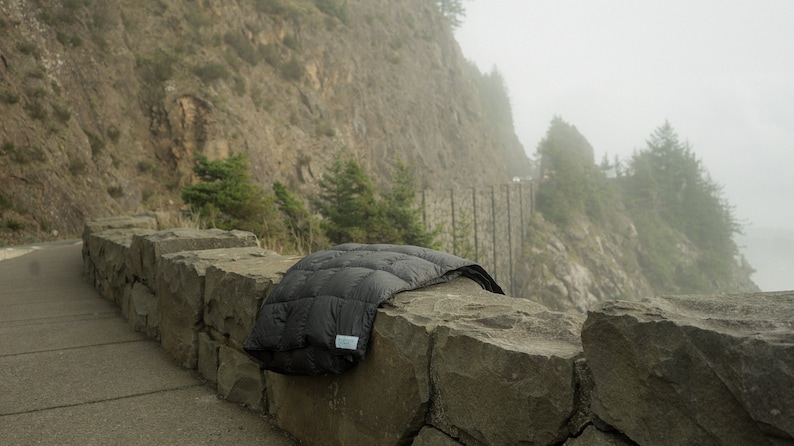 May include: A black down comforter is draped over a stone wall. The wall is on the edge of a road with a steep cliff in the background. The scene is foggy and misty.