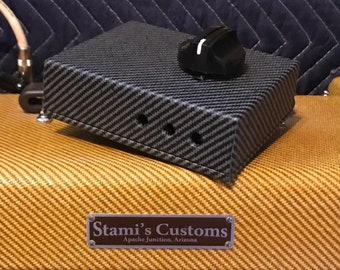 Puede incluir: Un pedal de guitarra negro y gris con una sola perilla. El pedal está sobre una superficie de tweed amarilla y marrón. El texto "Stami's Customs" y "Apache Junction, Arizona" está en una placa plateada sobre la superficie.