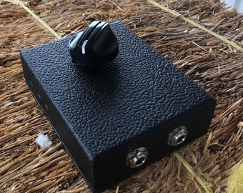 Puede incluir: Una caja negra con una sola perilla y dos conectores de 6,35 mm. La caja tiene una superficie texturizada.
