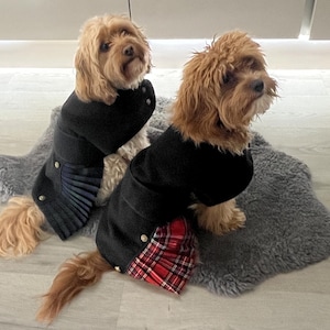 May include: Two small, light brown, fluffy dogs wearing black coats. One coat features a dark blue pleated skirt detail, and the other coat features a red and black plaid pleated skirt detail. Both coats are secured with small gold-toned buttons.