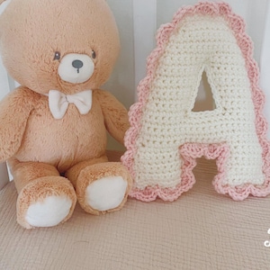 May include: A white crocheted letter A with a pink scalloped edge. The letter is sitting on a pink and white crib sheet next to a brown teddy bear with a white bow tie.