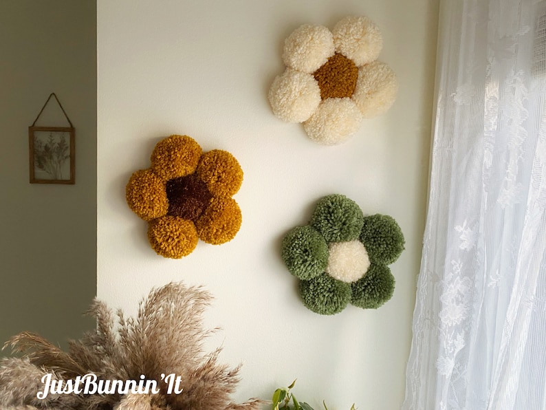 May include: Three handmade pom-pom flower wall decorations in yellow, white, and green. The flowers are made of yarn and have a center circle of a different color.