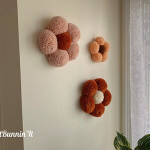 Op de afbeelding: Drie handgemaakte viltbloemen wanddecoraties in roze en bruine tinten. De grootste bloem is roze met een bruin centrum, de middelste bloem is bruin met een wit centrum en de kleinste bloem is roze met een bruin centrum.
