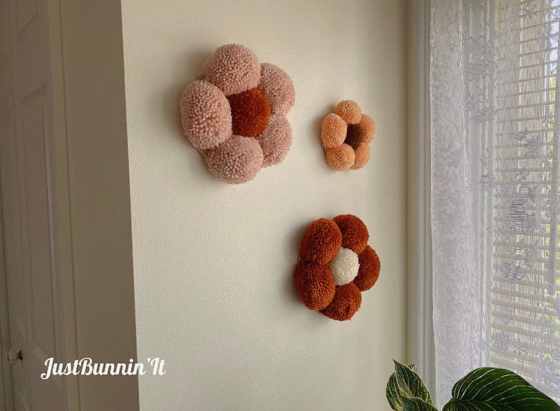 Op de afbeelding: Drie handgemaakte viltbloemen wanddecoraties in roze en bruine tinten. De grootste bloem is roze met een bruin centrum, de middelste bloem is bruin met een wit centrum en de kleinste bloem is roze met een bruin centrum.