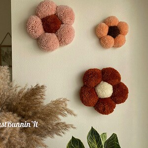 Op de afbeelding: Drie handgemaakte viltbloemen wanddecoraties in roze, bruin en wit. De bloemen zijn gemaakt van pompoenen en hebben een eenvoudig, modern ontwerp.