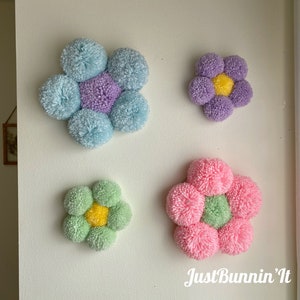May include: Four colorful pom-pom flower wall decorations in shades of blue, purple, green, and pink. The flowers are arranged in a staggered pattern on a white wall. The text "Just Bunnin' It" is visible in the bottom right corner of the image.