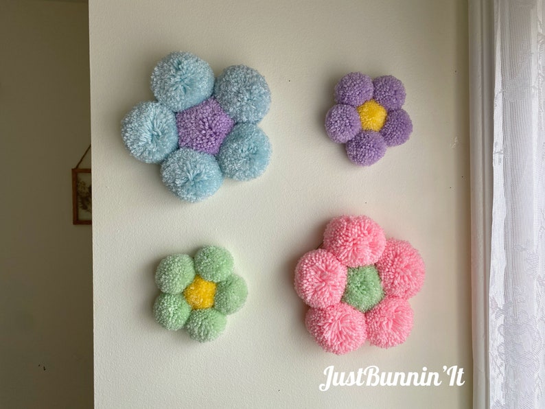 May include: Four colorful pom-pom flower wall decorations in shades of blue, purple, green, and pink. The flowers are arranged in a staggered pattern on a white wall. The text "Just Bunnin' It" is visible in the bottom right corner of the image.